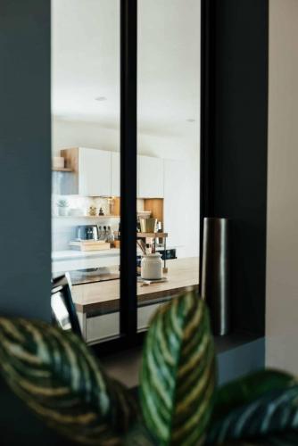 Vue sur la cuisine blanche et bois à travers une verrière intérieure noire intégrée à un mur vert foncé.