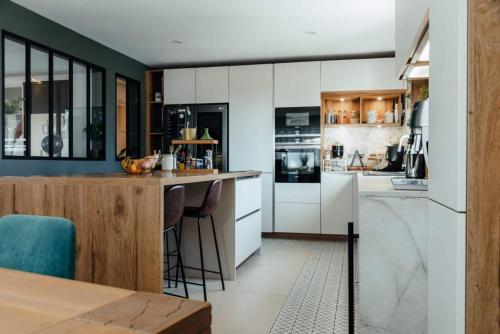Cuisine rénovée blanche et bois avec grand îlot central, espace repas et tabourets hauts noirs.